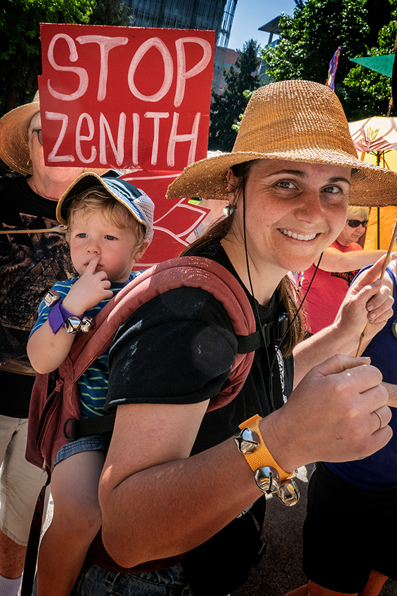 A smiling mother carrying her small toddler on her back. Behind her is a sign that says "Stop Zenith".