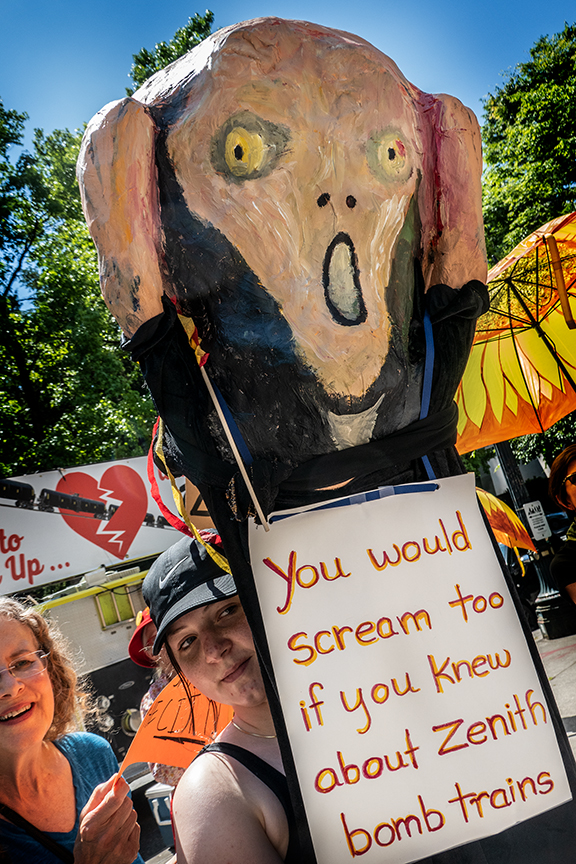 Activistas sosteniendo un cartel con la parte superior de una réplica de la pintura de Edvard Munch "El Grito". El texto dice "Tú también gritarías si supieras acerca de los trenes Zenith Bomb".