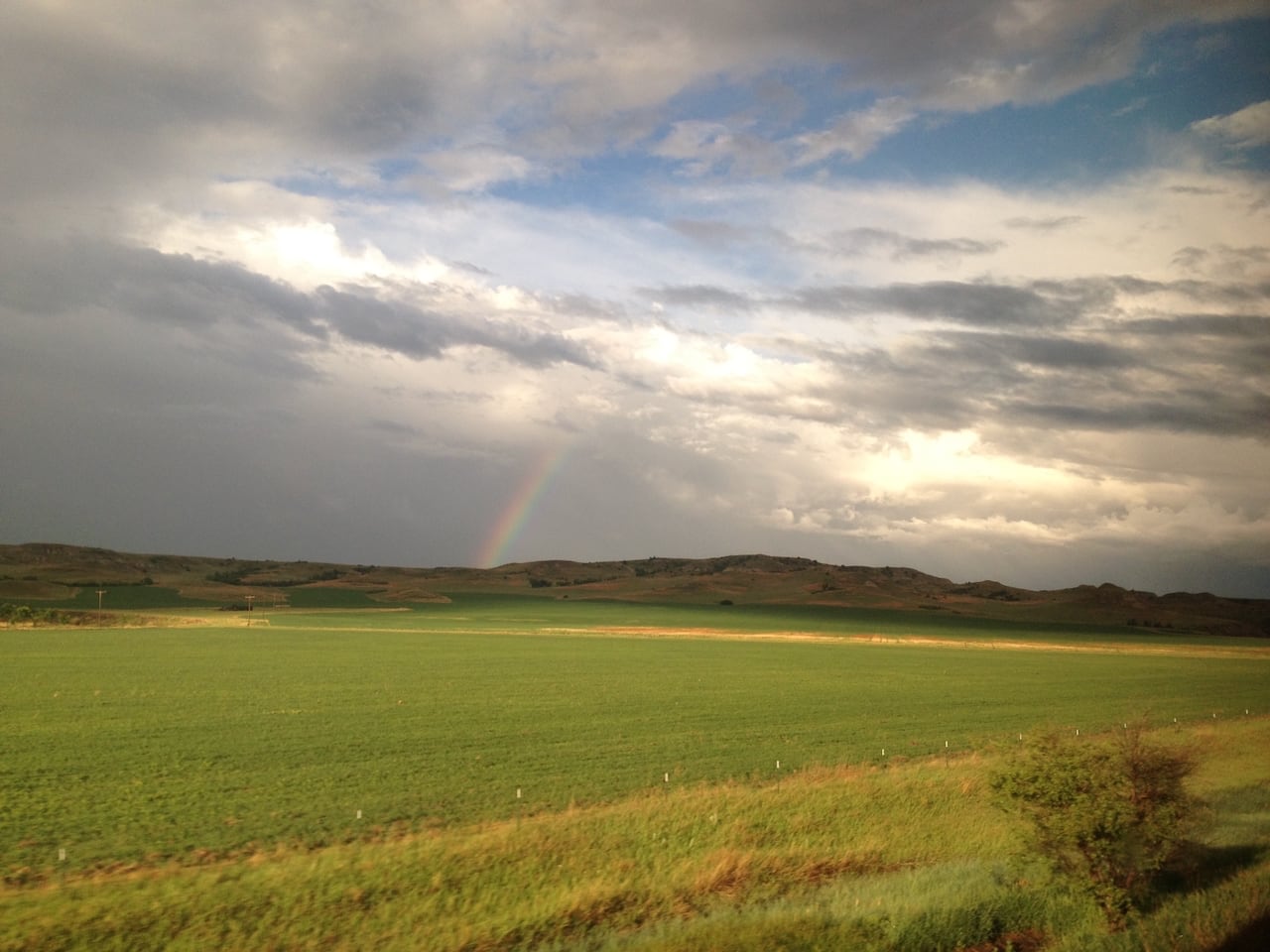 Una vista de un campo verde con colinas bajas en la distancia. El cielo en la distancia es oscuro y gris pero hay una mancha azul arriba. Y hay un arco iris.
