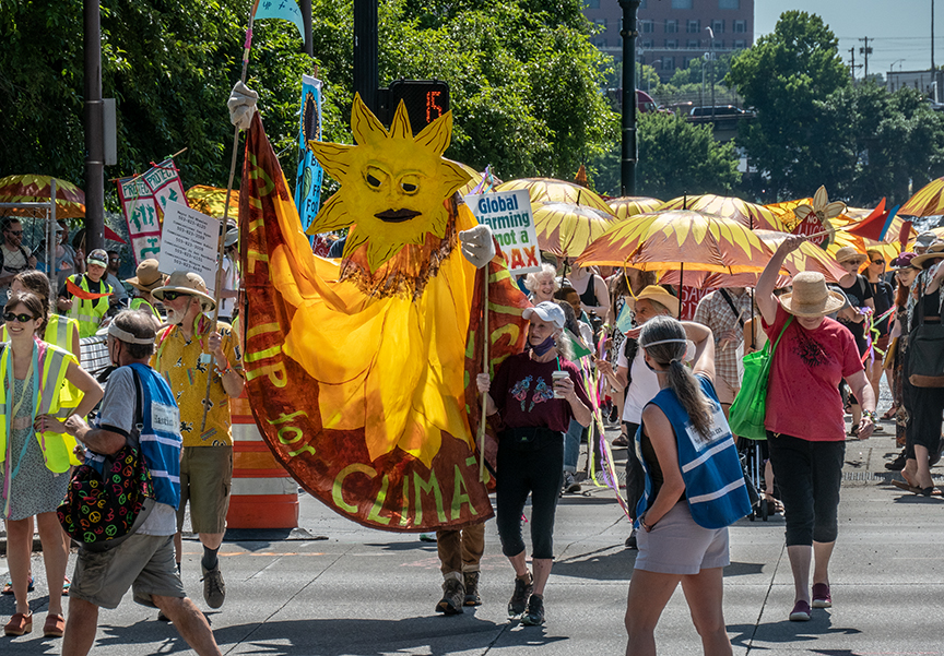 People marching in the Stop Zenith Parade. There is a "sun" puppet in the foreground.