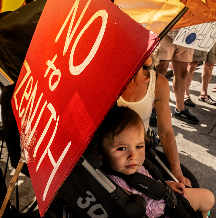 A child in a stroller getting some shade from the sun under a sign that says "Say No to Zenith".